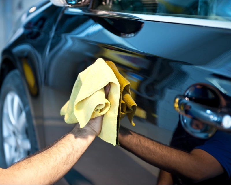 Person polishing a black car with a yellow cloth, reflecting nearby surroundings.