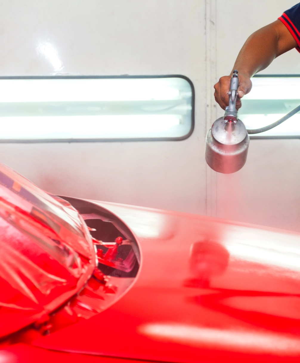 Person spray painting a red car in a workshop, with a bright light in the background.