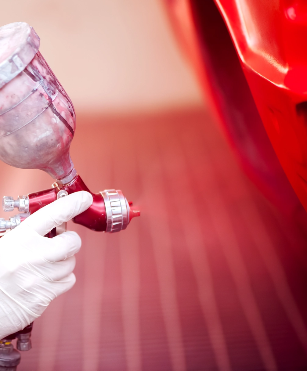 Close-up of a person spray painting a red car with a paint gun, wearing a white glove.