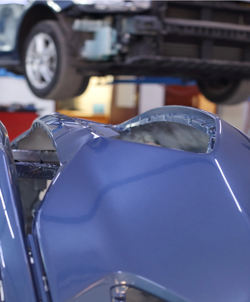 A blue car bumper is detached and visible in the foreground, with a partially disassembled car on a lift in the background.