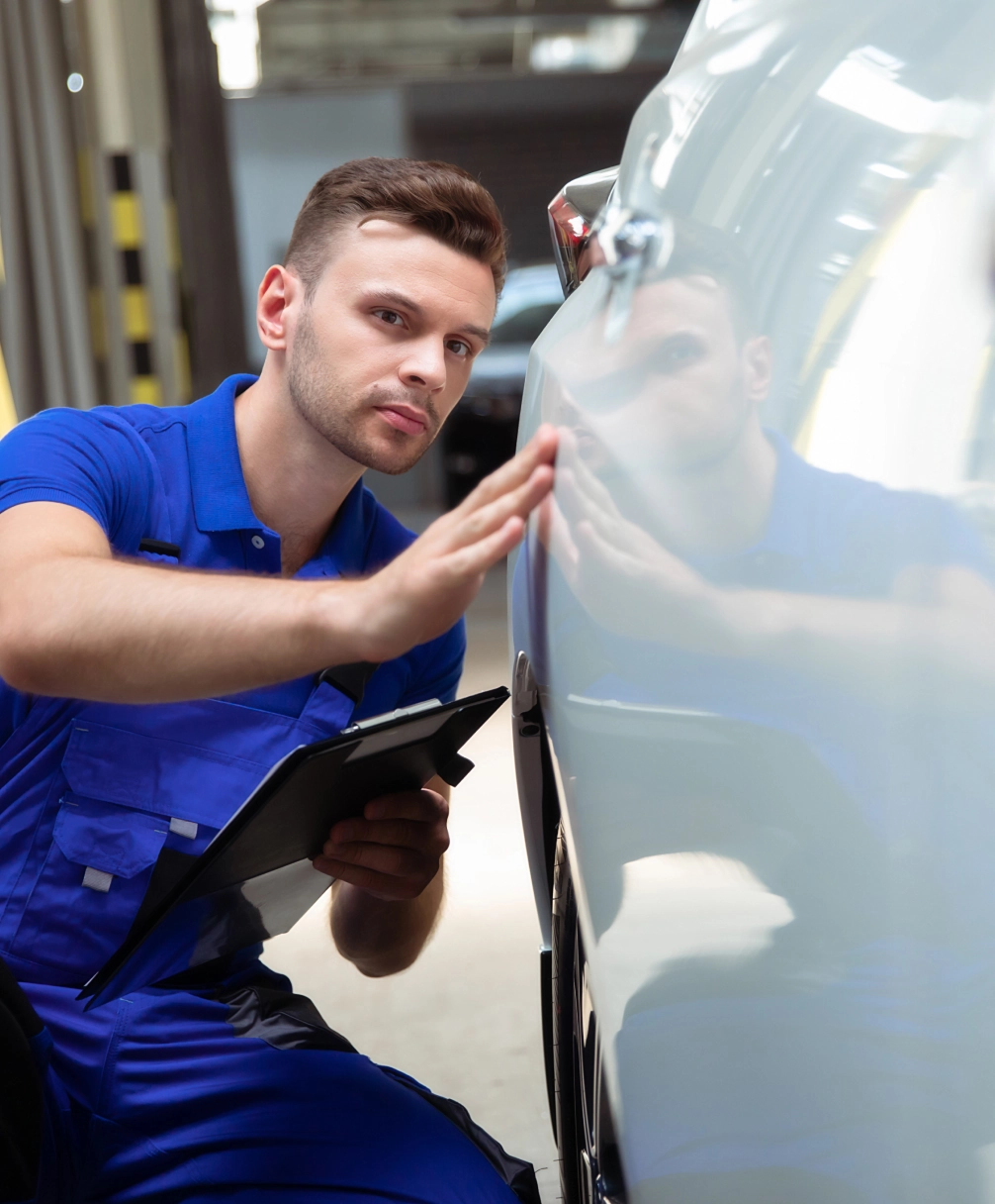 A mechanic in a blue uniform inspects a car's exterior while holding a clipboard.