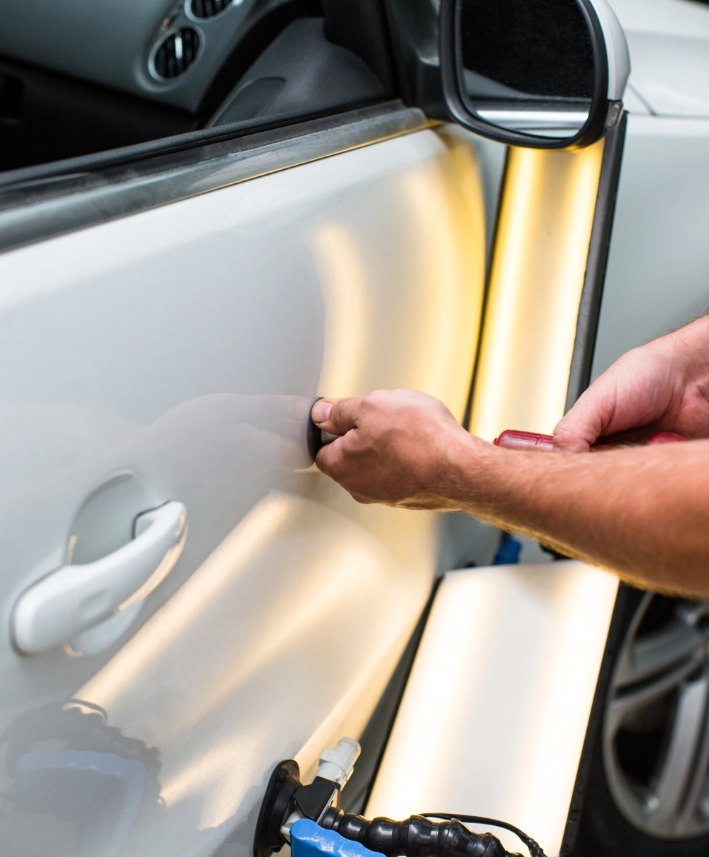 Person repairing a dent on a car door using a tool, with a light reflection assisting in visualizing the dent.