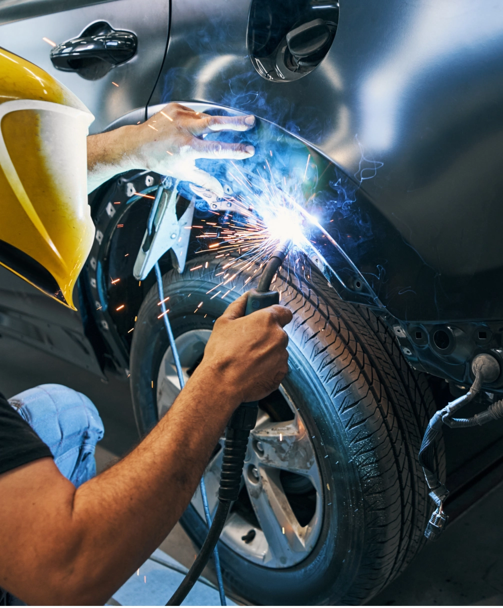 A person wearing a yellow helmet welds the fender of a car, creating bright sparks.