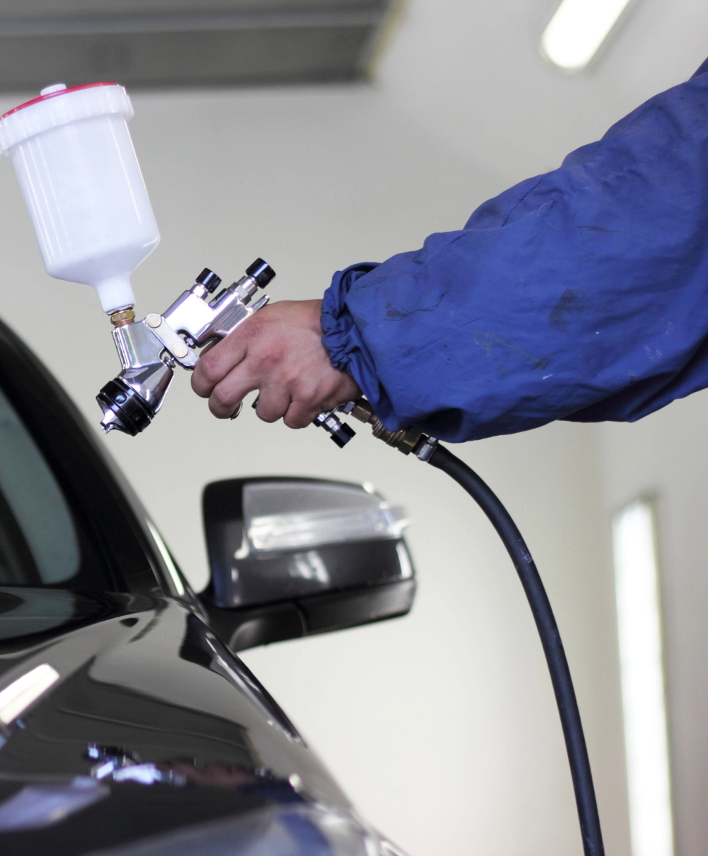 Person in a blue jacket using a spray gun to paint a black car in a garage.