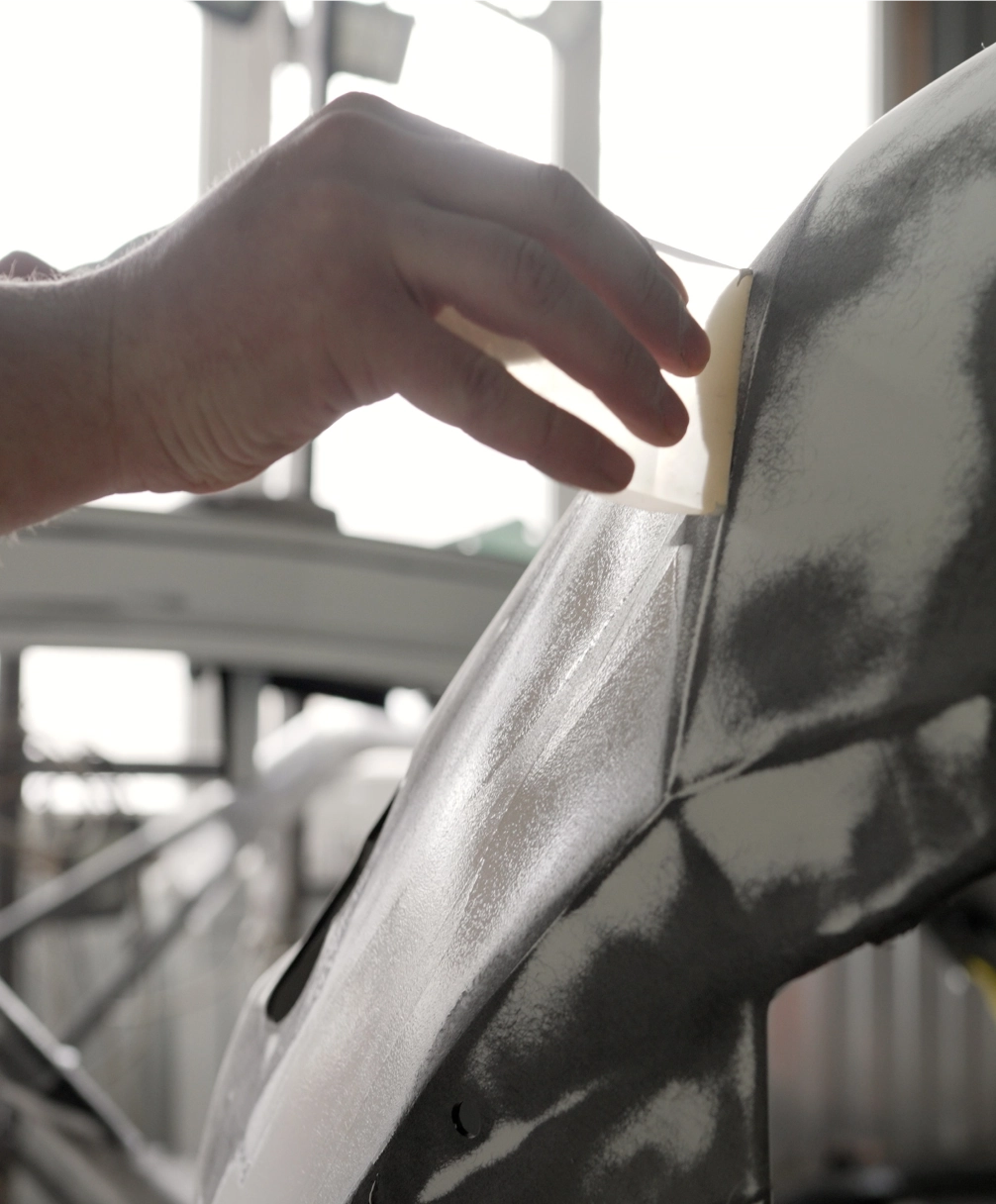 A hand applies a piece of tape to a metallic car part in a bright workshop.