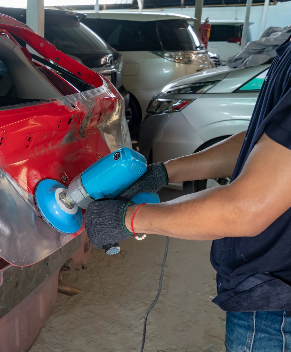 Person using a blue electric buffer on a red car's body in a workshop setting.